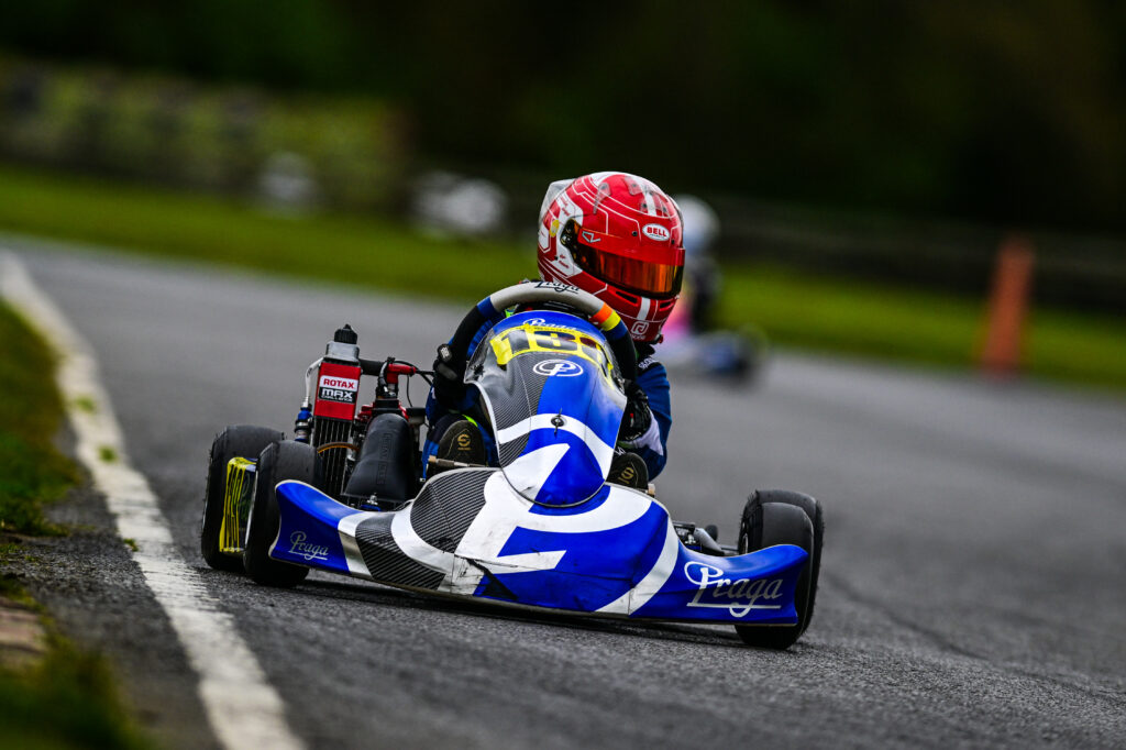 About RJ Racing 39. A young kart racer in a blue and white Praga kart, wearing a red helmet, races on an outdoor track with another racer in the distant background.