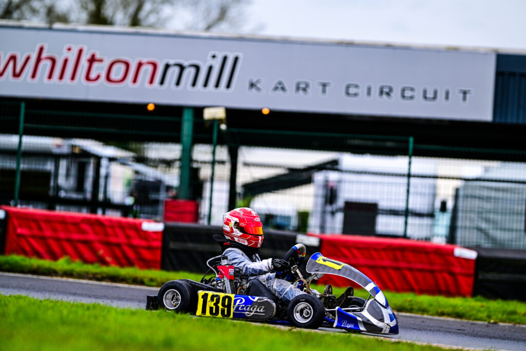 About RJ Racing 39. A young kart racer in a blue and white Praga kart, wearing a red helmet, races at Whilton Mill Kart Circuit. The track's sign is visible in the background.